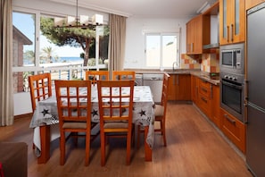 A dining area with a table and chairs, a kitchen with wooden cabinets and appliances, and a view of the sea through large windows.