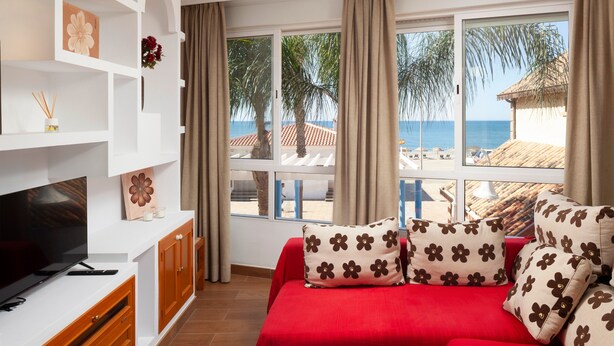 Une pièce avec une télévision, un canapé rouge orné de coussins à motifs floraux, une table blanche et une vue sur la plage.