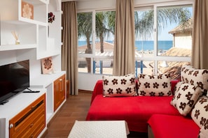 A room with a TV, a red sofa with flower-patterned pillows, a white table, and a view of the beach.