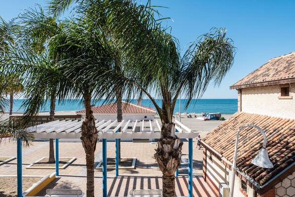 A beachside view with palm trees, a pergola, and a building with a tiled roof.