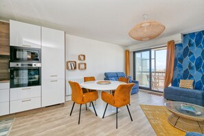 Uma cozinha moderna com eletrodomésticos embutidos, uma sala de jantar com cadeiras laranja, um sofá azul e uma janela grande.
