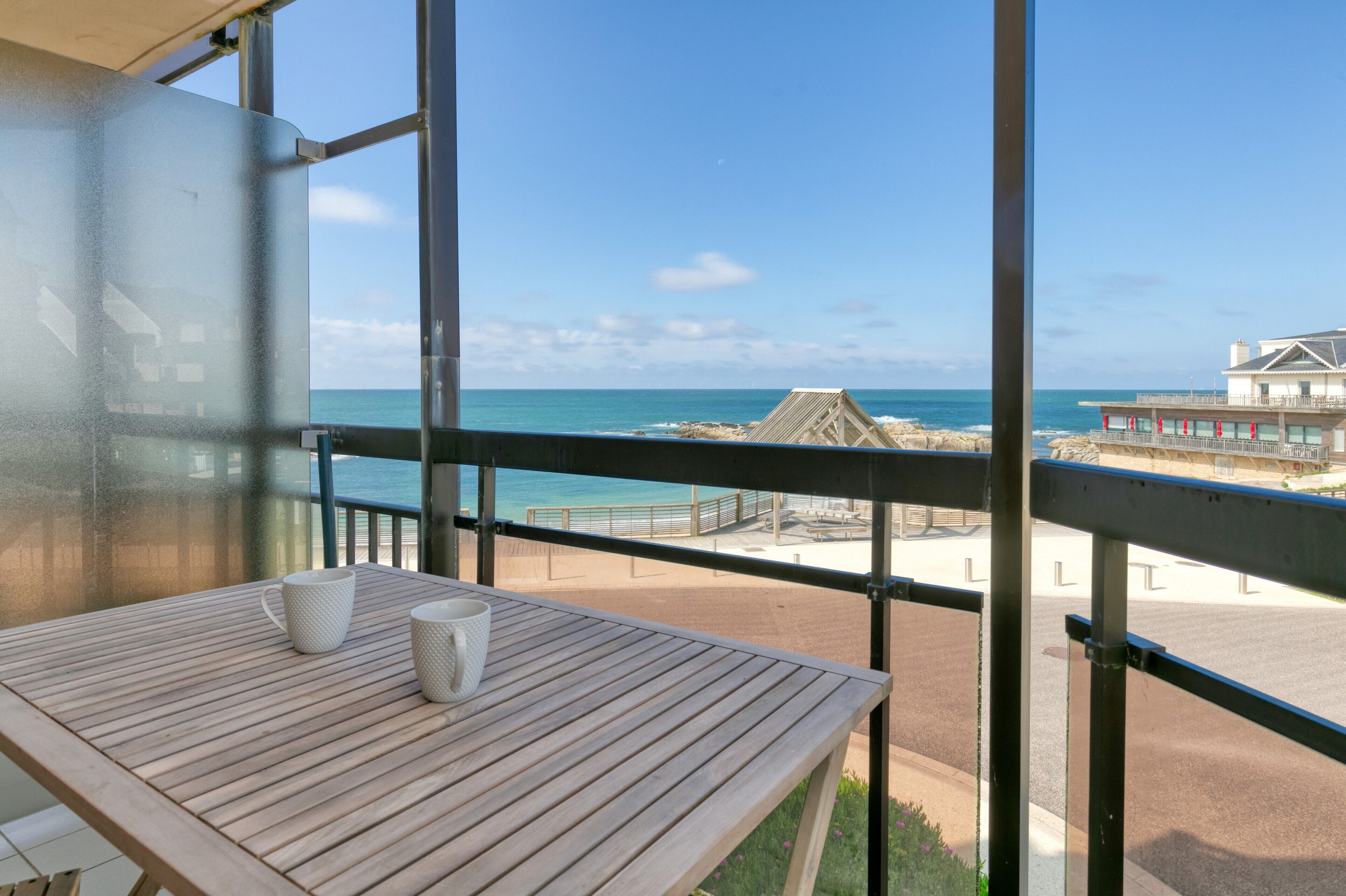 Varanda com mesa de madeira e duas canecas brancas, com vista para uma praia que inclui uma estrutura de palha e um edifício com varandas.