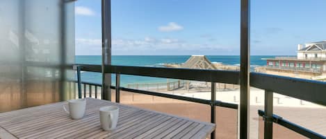 Varanda com mesa de madeira e duas canecas brancas, com vista para uma praia que inclui uma estrutura de palha e um edifício com varandas.