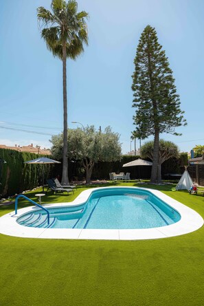 Ein Swimmingpool mit einer umlaufenden Rasenfläche, Liegestühlen und einem hohen Baum.