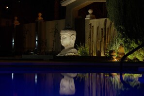 A serene nighttime garden with a large Buddha statue, bamboo plants, and a reflective pool.