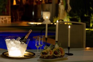A table with a glass bowl of ice, two glasses, a bottle of champagne, and a fruit platter, with candles and a pool in the background.
