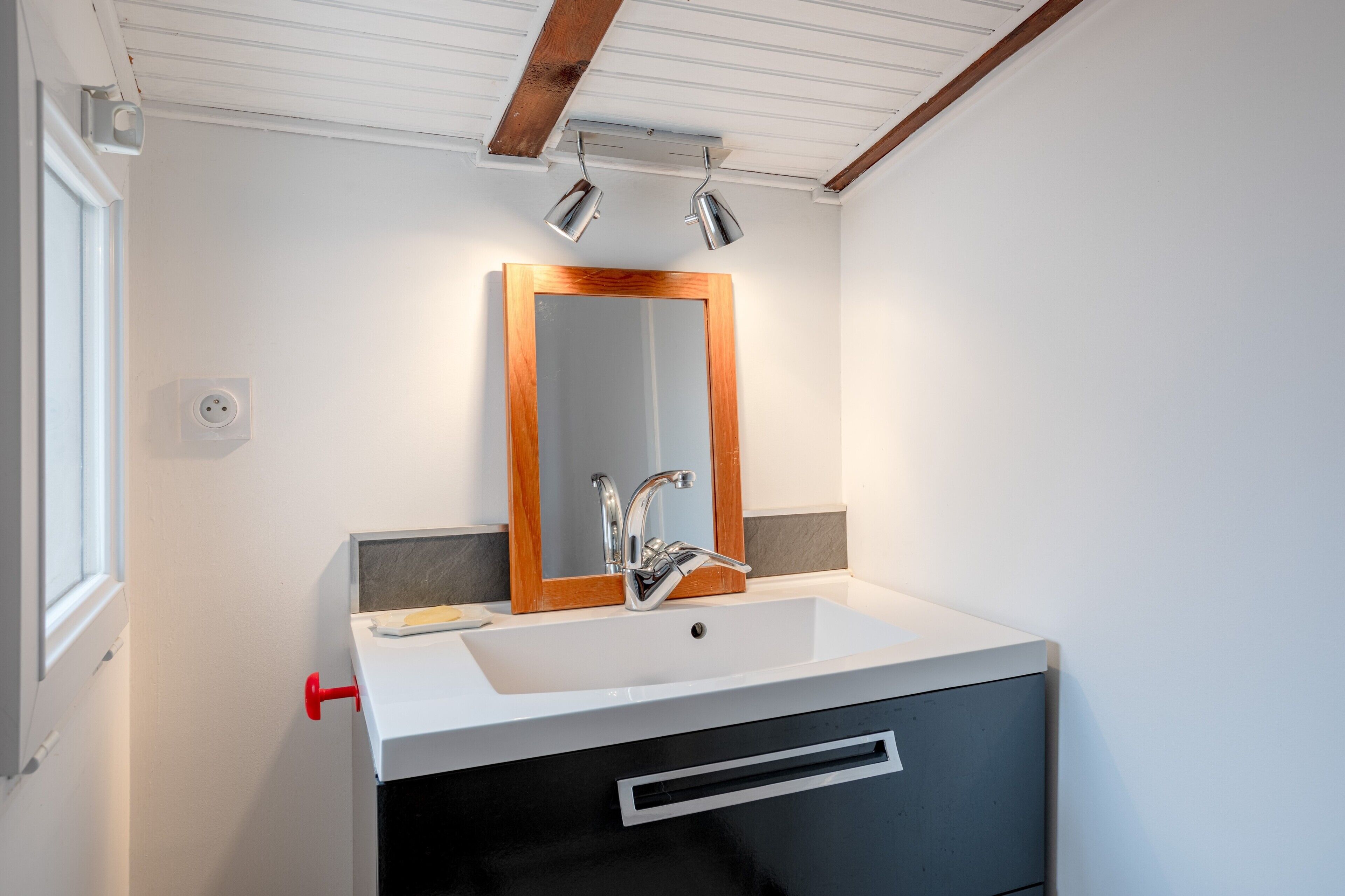 Un bagno con un lavandino bianco, uno specchio con cornice in legno e una maniglia di porta rossa.