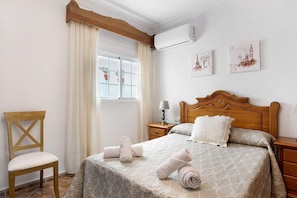 Una cama bien hecha con toallas enrolladas, una silla de madera, una mesita de noche con lámpara y una ventana con cortinas.