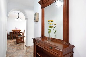 Un pasillo con una consola de madera, un espejo y un florero con flores. Se observa un comedor con una mesa y sillas al fondo.