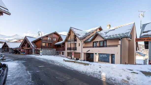 A snow-covered street with wooden buildings, including a hotel with a sign 'Office de location.' - Apartment 'La Croix Du Sud' with Mountain View and Balcony (Valmeinier)