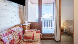 A cozy room with a wooden wall, a bed with patterned bedding, a small wooden nightstand, and a view of a balcony with a railing. - Apartment 'La Croix Du Sud' with Mountain View and Balcony (Valmeinier)