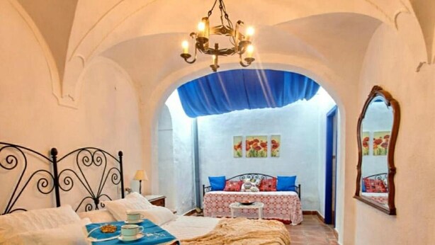 Une chambre à coucher avec un lit, une table recouverte d’une nappe bleue, un lustre et un miroir au mur.