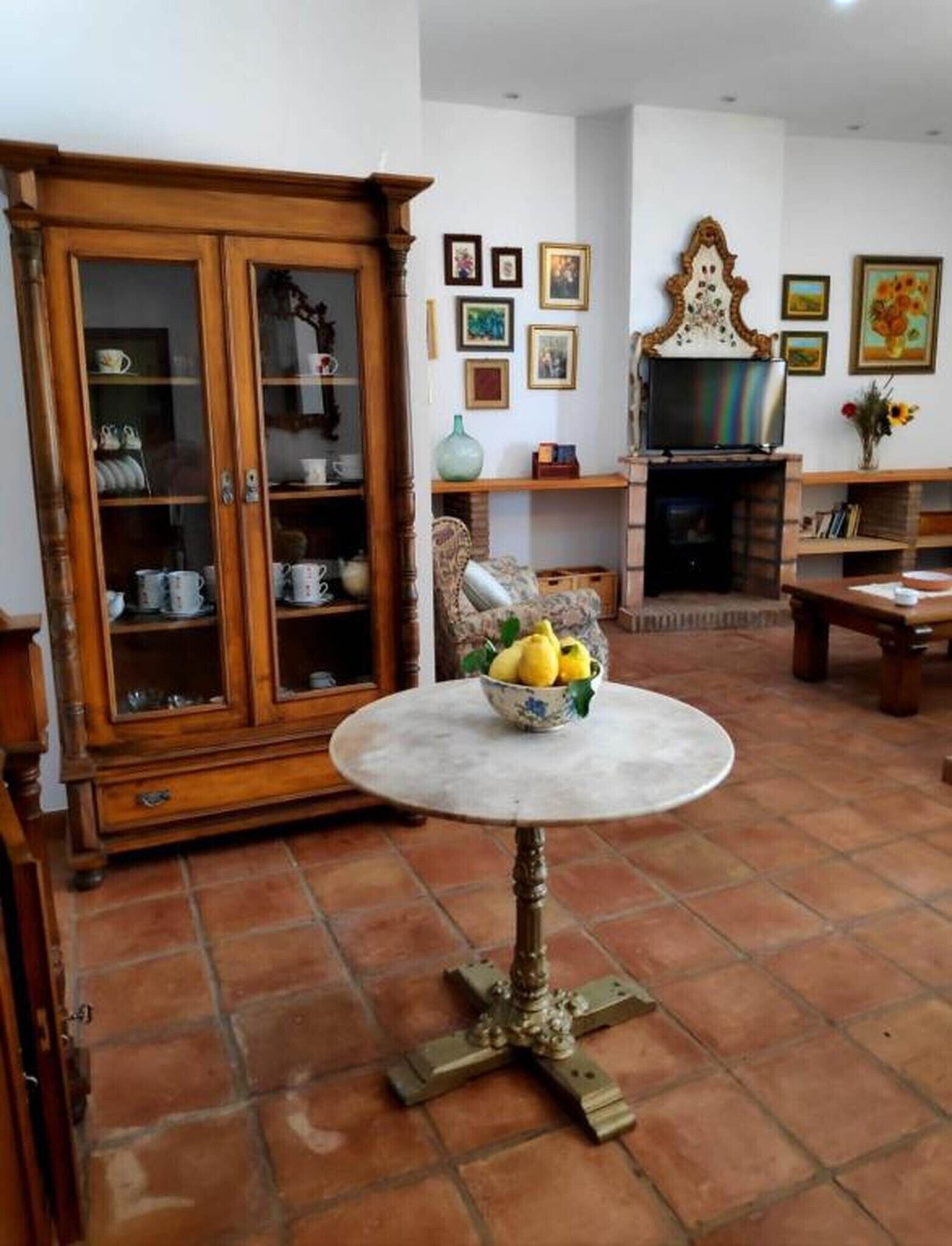 A living room with a fireplace, a wooden cabinet with glass doors, a round table with a bowl of fruit, and a tiled floor.