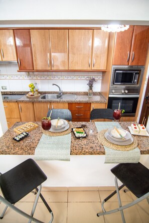 Una cocina con armarios de madera, un microondas y un horno. Hay una mesa de comedor con sillas y una pequeña isla con un jarrón y un frutero.