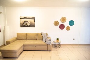Una sala de estar con un sofá seccional color beige, una mesita auxiliar con una planta, un cuadro colgado en la pared de un barco y decoración mural circular.