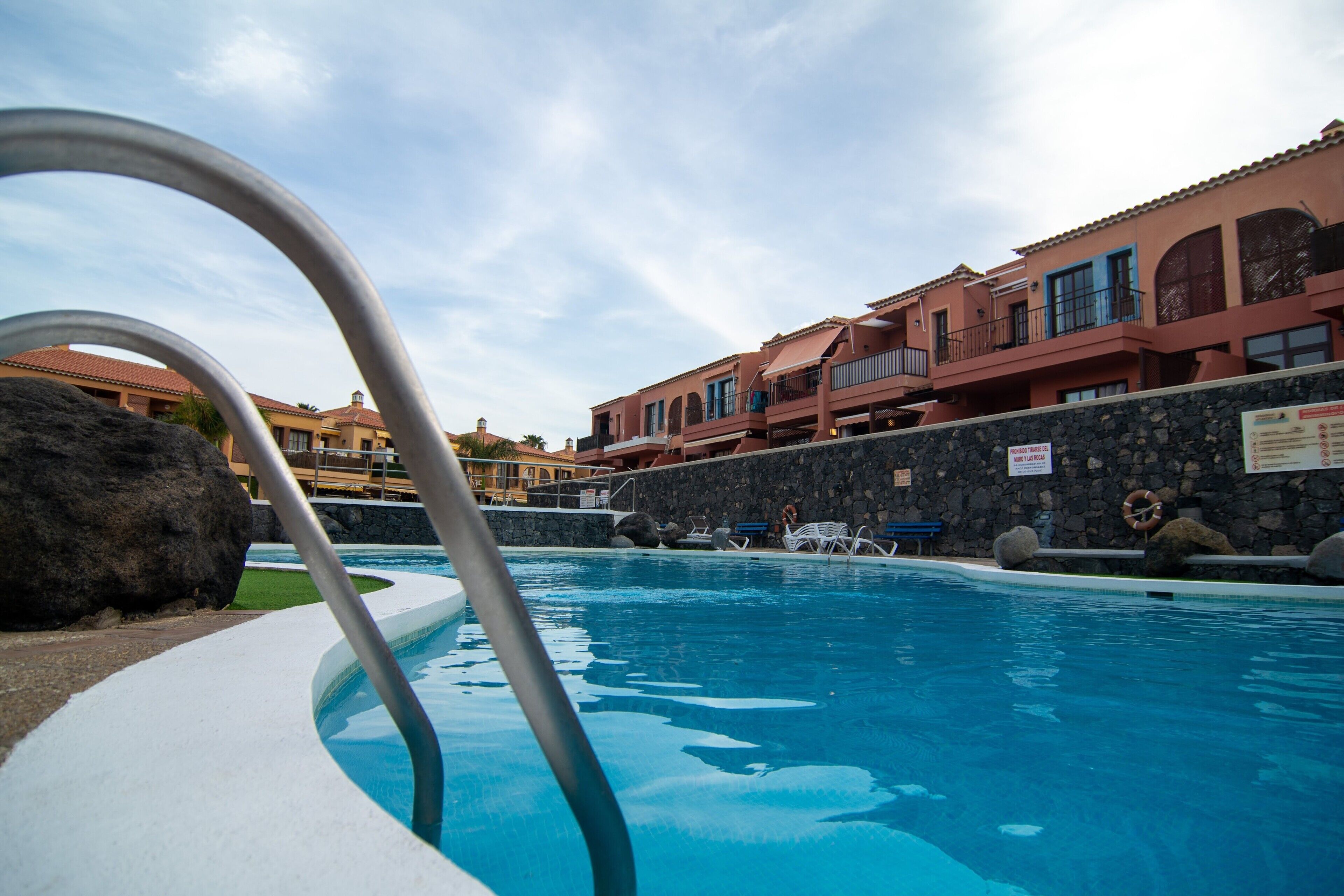 A swimming pool with a metal ladder, stone walls, and a view of hotel buildings with balconies.