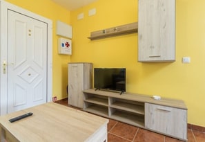 A room with a yellow wall, a white door, a TV on a wooden stand, a remote control on a table, and a mounted emergency medical kit.
