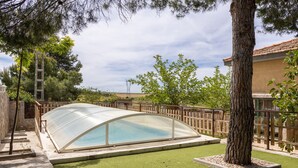 Una piscina con cerramiento de vidrio, rodeada por una valla de madera, árboles y un sendero de piedra.