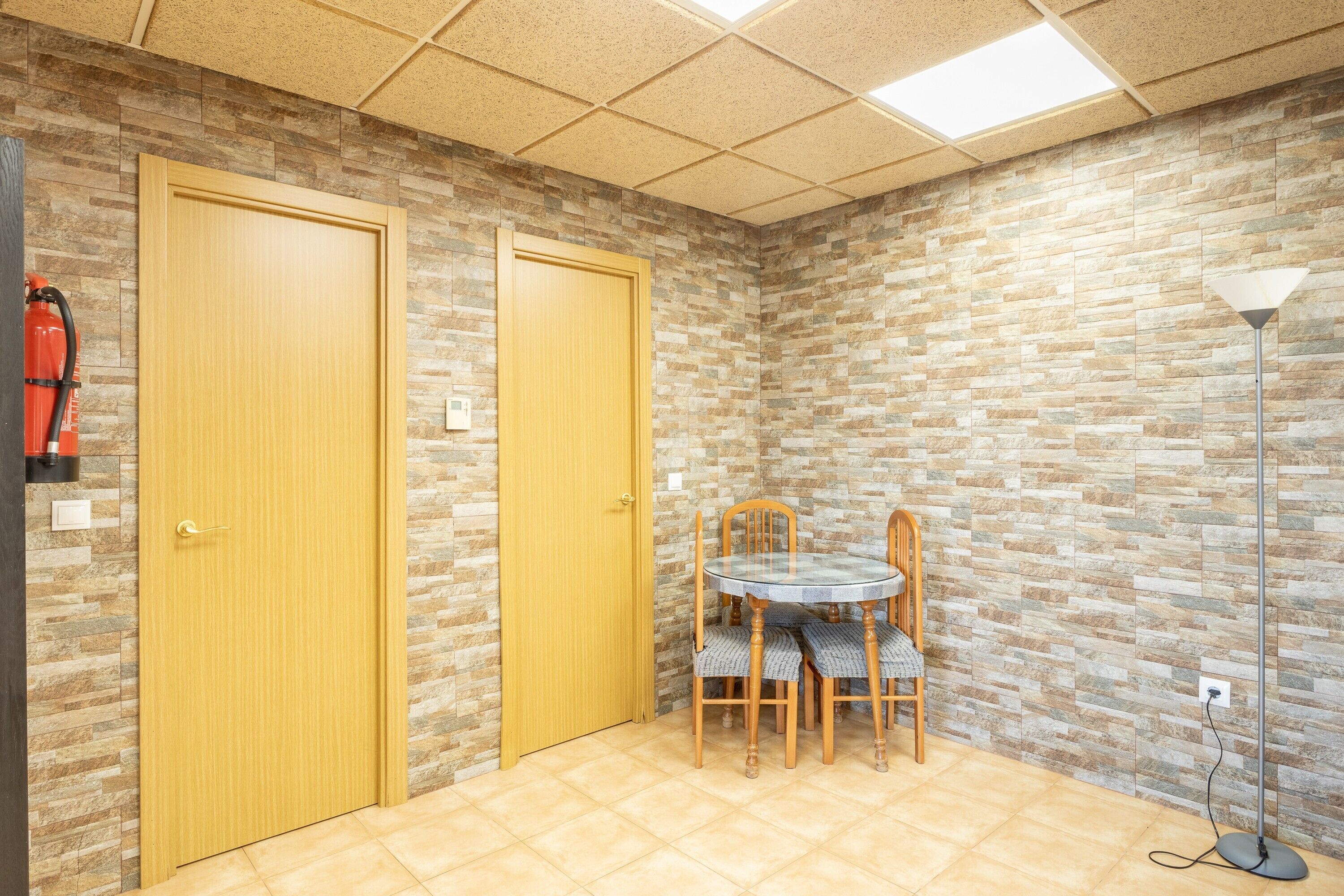 Een kamer met twee houten deuren, een ronde tafel met stoelen, een vloerlamp en een brandblusser aan de muur.