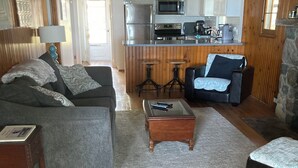 Un salon confortable avec un canapé, une table basse et une cheminée. On aperçoit, au fond, une cuisine équipée d’un four à micro-ondes et d’un réfrigérateur.