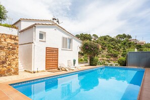 A white villa with a swimming pool, stone wall, and a view of a hill with trees.