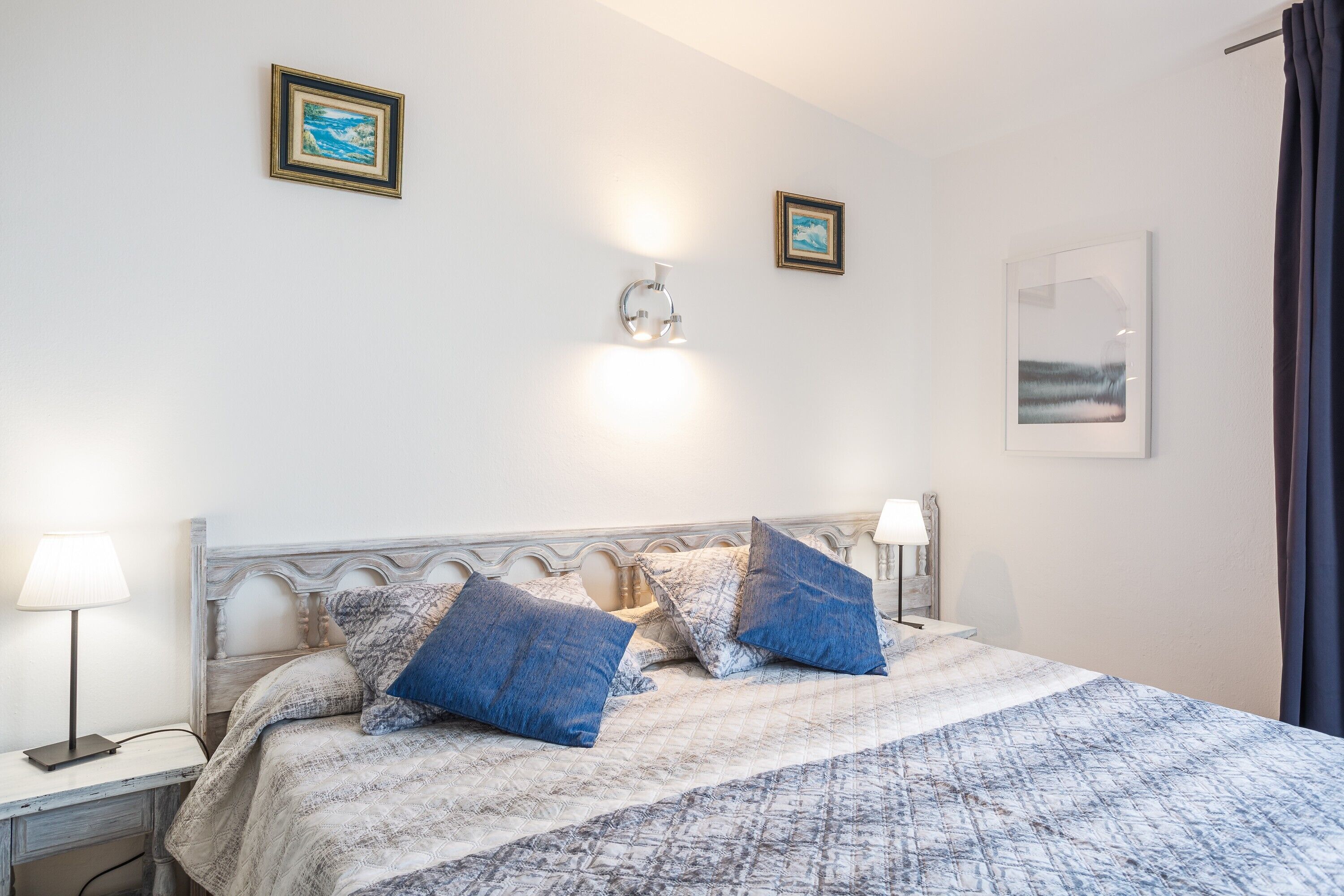 Een slaapkamer met een bed, twee bedlampen, een wandlamp en twee ingelijste foto's.
