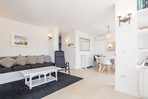 A modern living room with a sofa, a dining area with a table and chairs, and a kitchen in the background.
