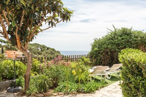 A garden with a view of the sea, a wooden fence, and a row of white sun loungers.