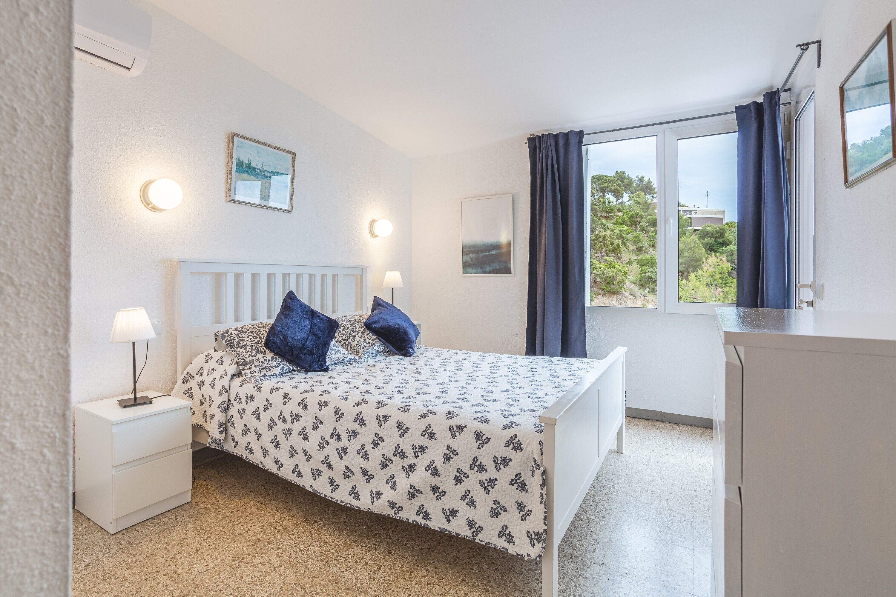 Une chambre à coucher avec un lit, des tables de chevet, une lampe, une fenêtre avec des rideaux et une vue sur l’extérieur.