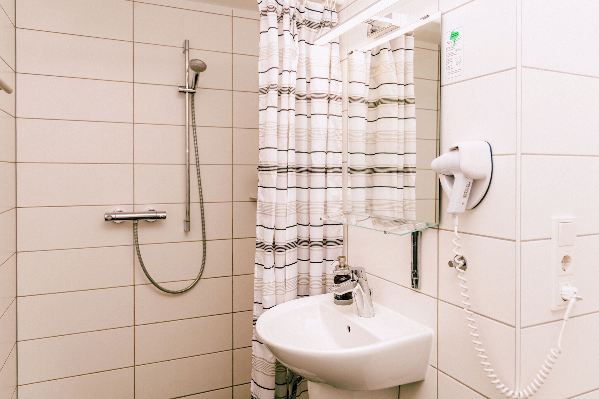 Un baño con ducha, lavamanos, secador de pelo y repisa de vidrio con artículos de aseo.