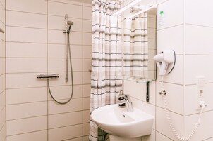 Un baño con ducha, lavamanos, secador de pelo y repisa de vidrio con artículos de aseo.