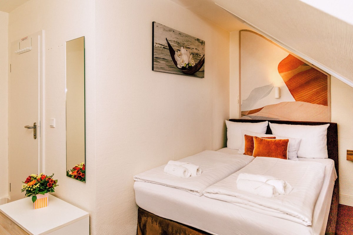 A hotel room with a bed, a bedside table with a vase of flowers, a large mirror, and a wall-mounted artwork.