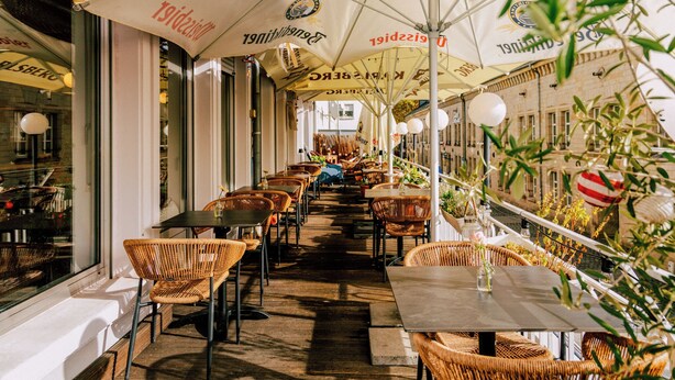 Un café al aire libre con sillas y mesas de mimbre bajo sombrillas grandes, con vistas a un edificio al fondo.