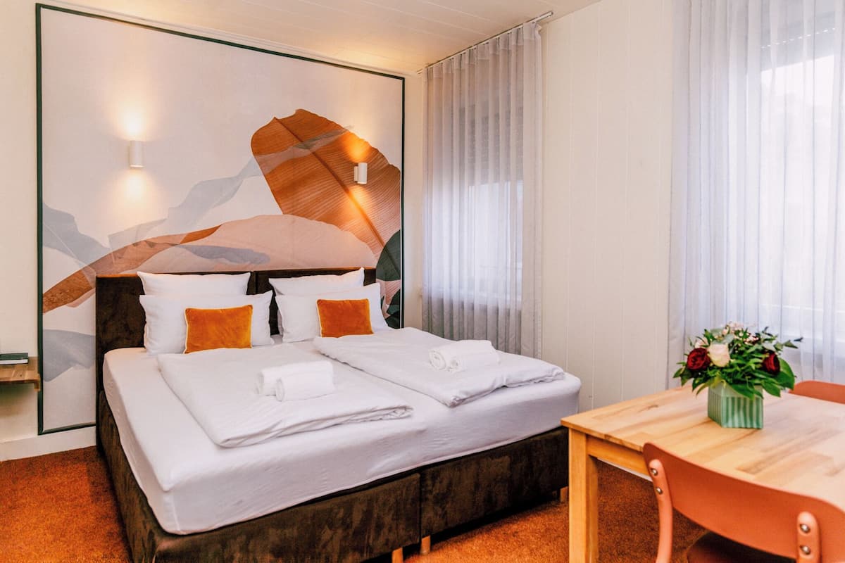 A neatly made bed with white linens and orange pillows, a wooden headboard, and a wall-mounted light. There is a wooden table with a vase of flowers and a chair to the right.