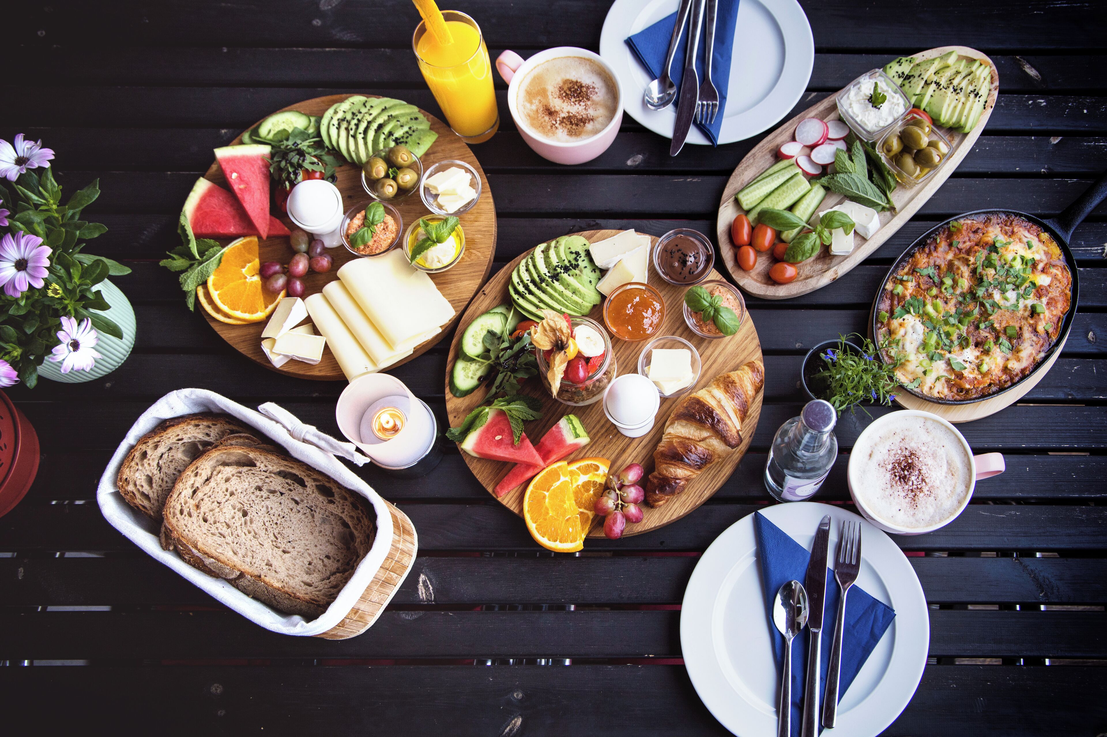 Verschiedene Speisen und Getränke auf einem Holztisch, darunter Brot, Käse, Obst und eine Schale mit Salat und Käse.