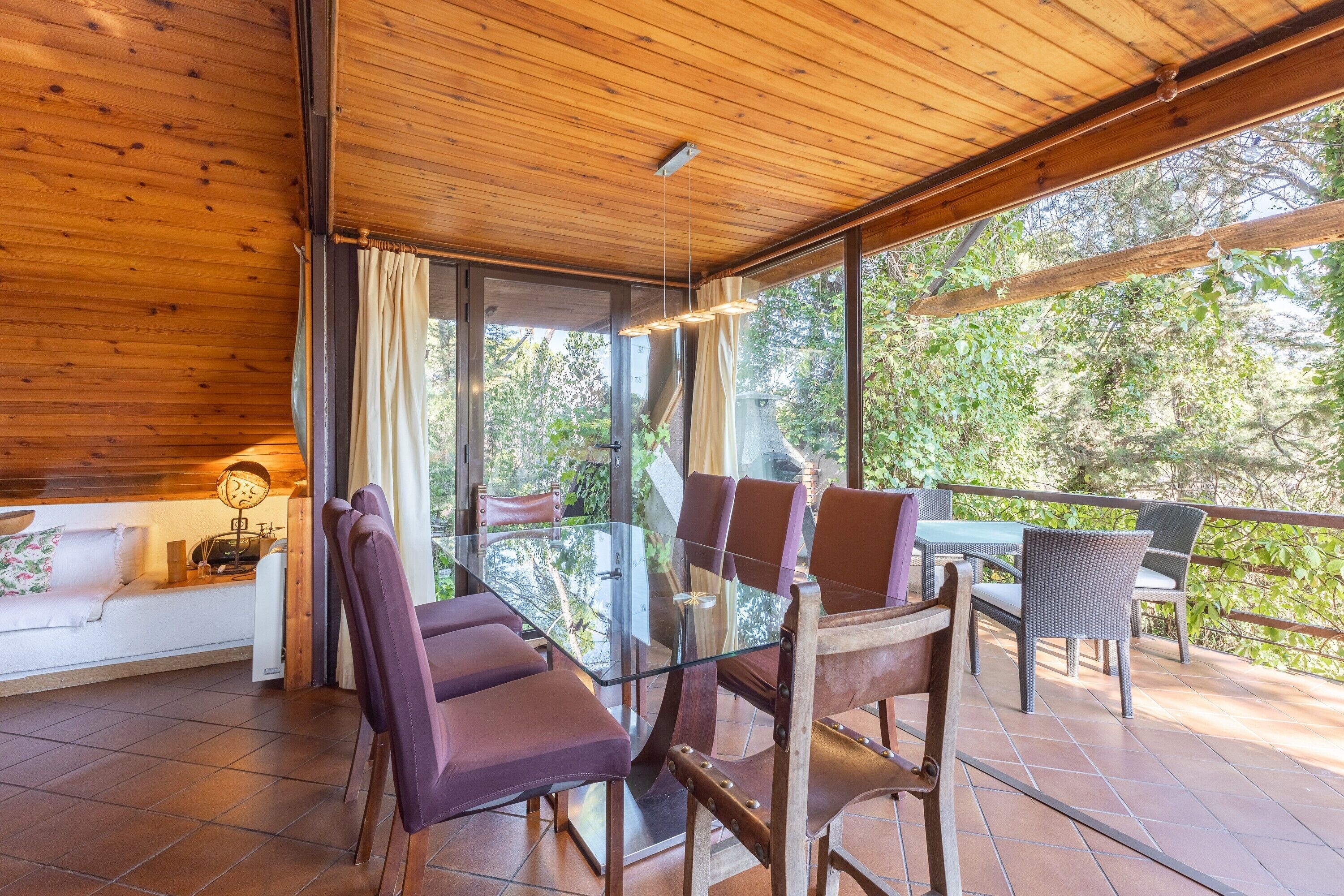 Soffitto in legno, tavolo da pranzo in vetro, sedie e un divano in una stanza con ampie finestre.