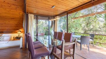 Soffitto in legno, tavolo da pranzo in vetro, sedie e un divano in una stanza con ampie finestre.