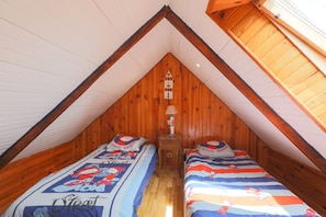 Een knus zolderkamertje met houten wanden en schuine plafonds, twee bedden met bijpassend beddengoed, een nachtkastje en een wandlamp.