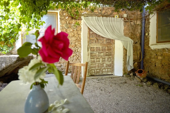 Un mur de pierres apparentes, une porte en bois avec un rideau, une table ornée d’un vase de fleurs et une chaise.