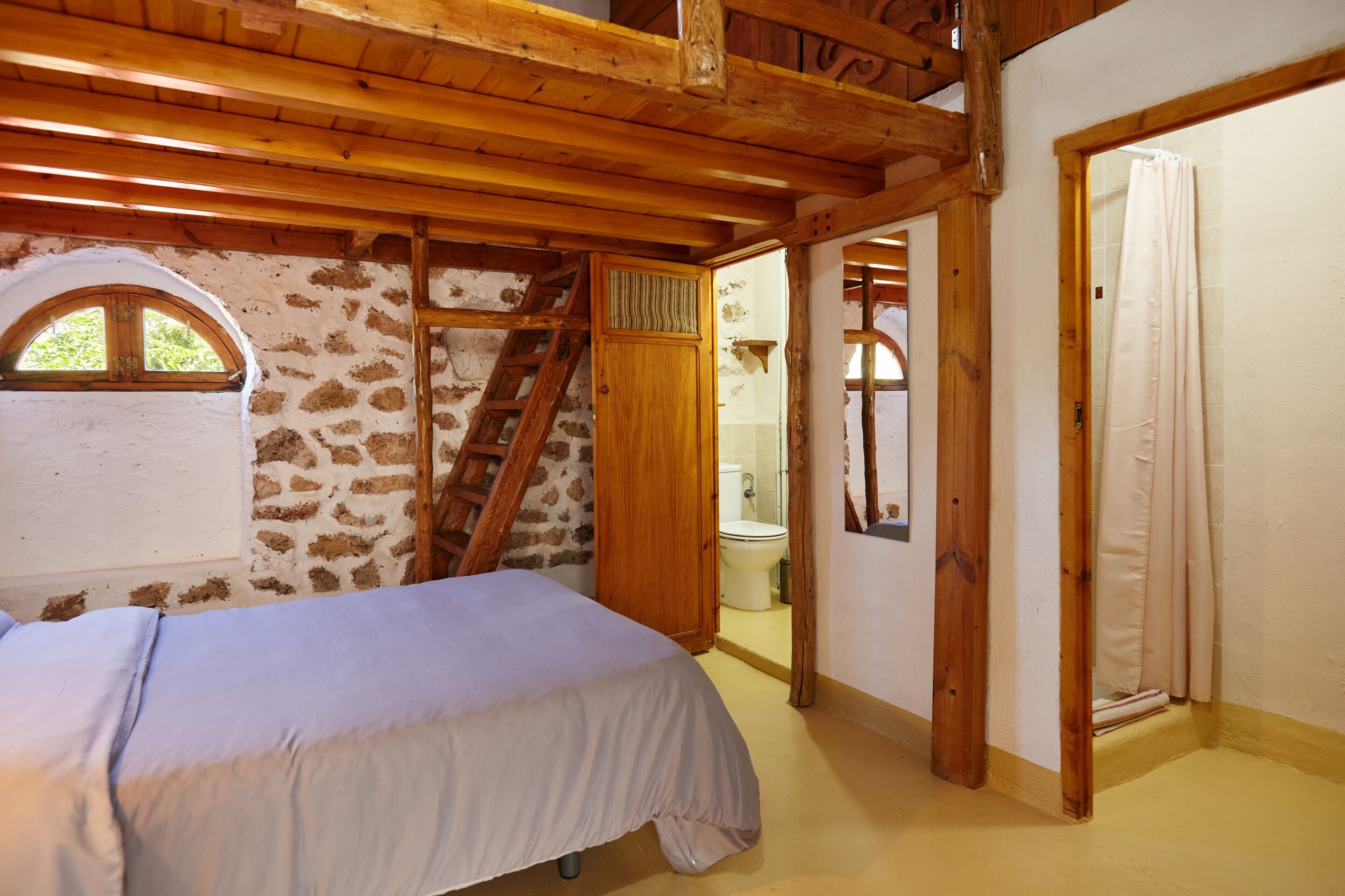 Un dormitorio con cama, vigas de madera, pared de piedra, ventana, baño con inodoro y lavabo, y una puerta de madera.
