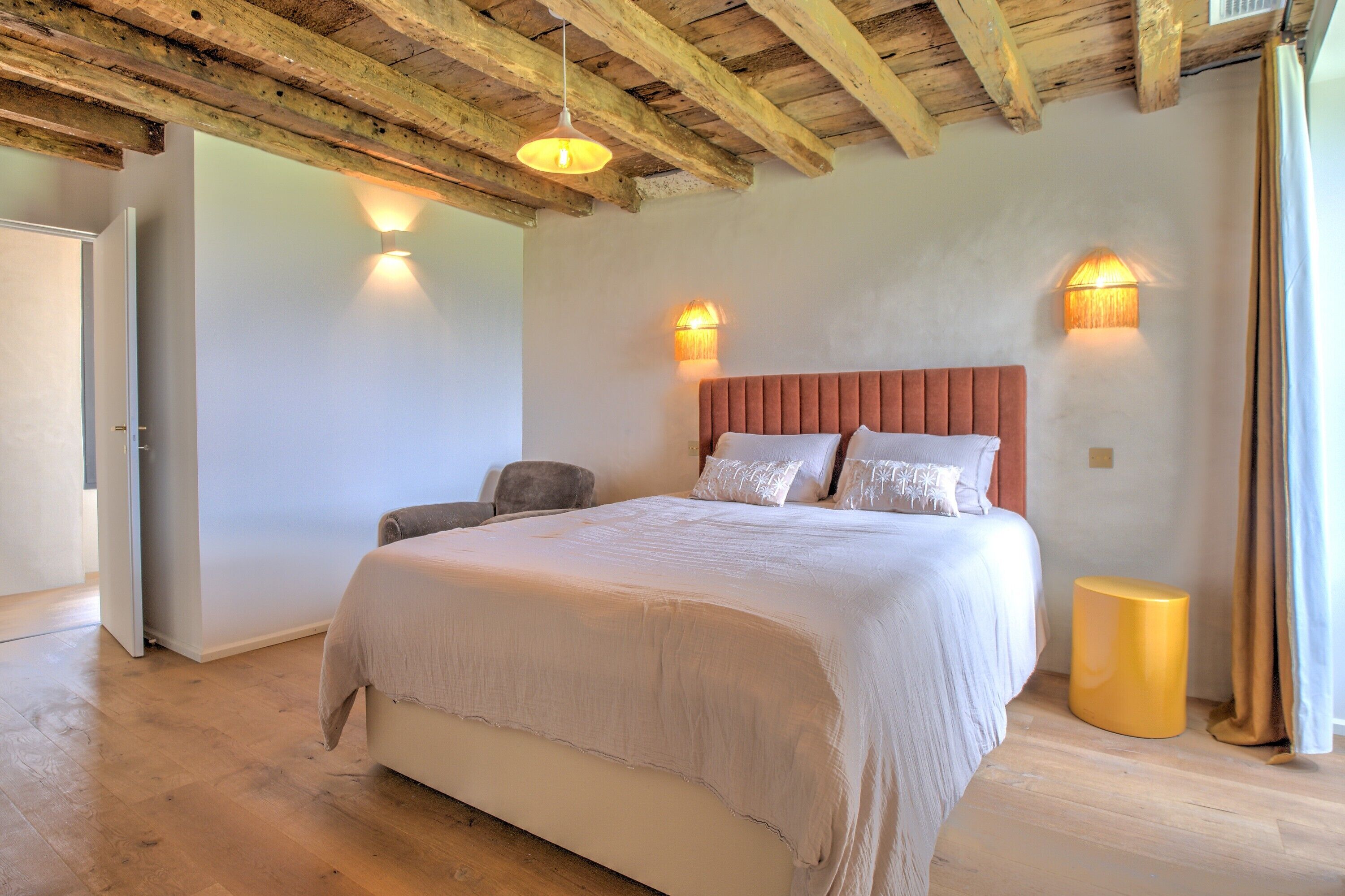 A bedroom with a large bed, wooden ceiling beams, two wall-mounted lights, and a small round stool.