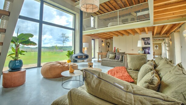 A spacious living area with a large window, a fireplace, and a high ceiling with exposed beams.