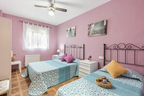 A bedroom with two beds, a ceiling fan, a window with curtains, a bedside table with a lamp, and artwork on the wall.