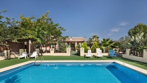 Una piscina rectangular con agua azul cristalina, rodeada de sillas de descanso blancas, una zona de estar techada y diversas plantas y árboles.