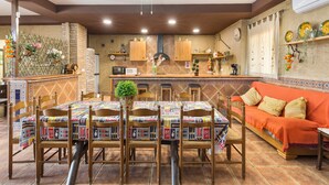 Comedor con mesa dispuesta para seis personas, sillas de madera alrededor, una planta en el centro y cocina al fondo.