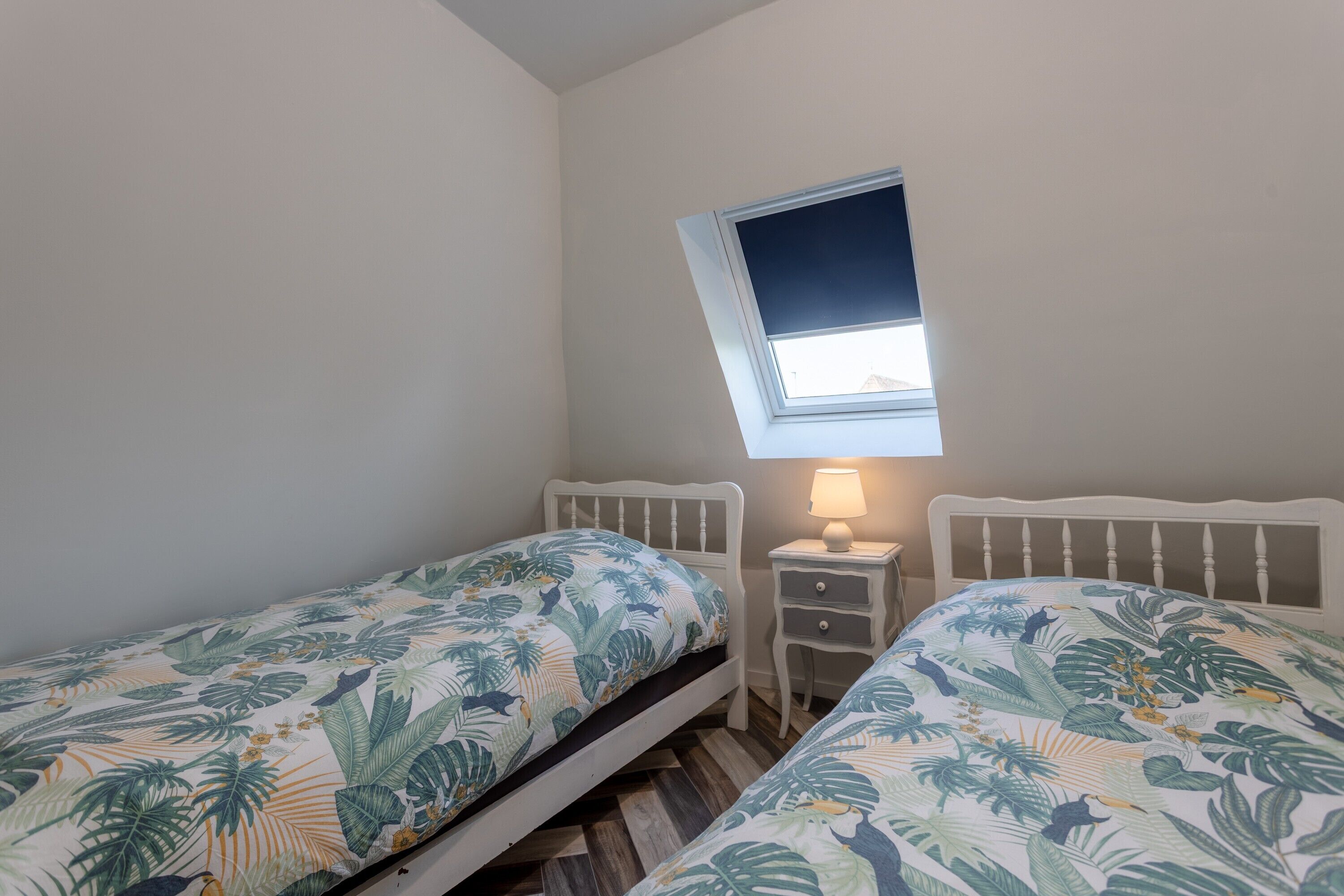 A bedroom with two single beds, a small bedside table with a lamp, and a window with a view of the sky.