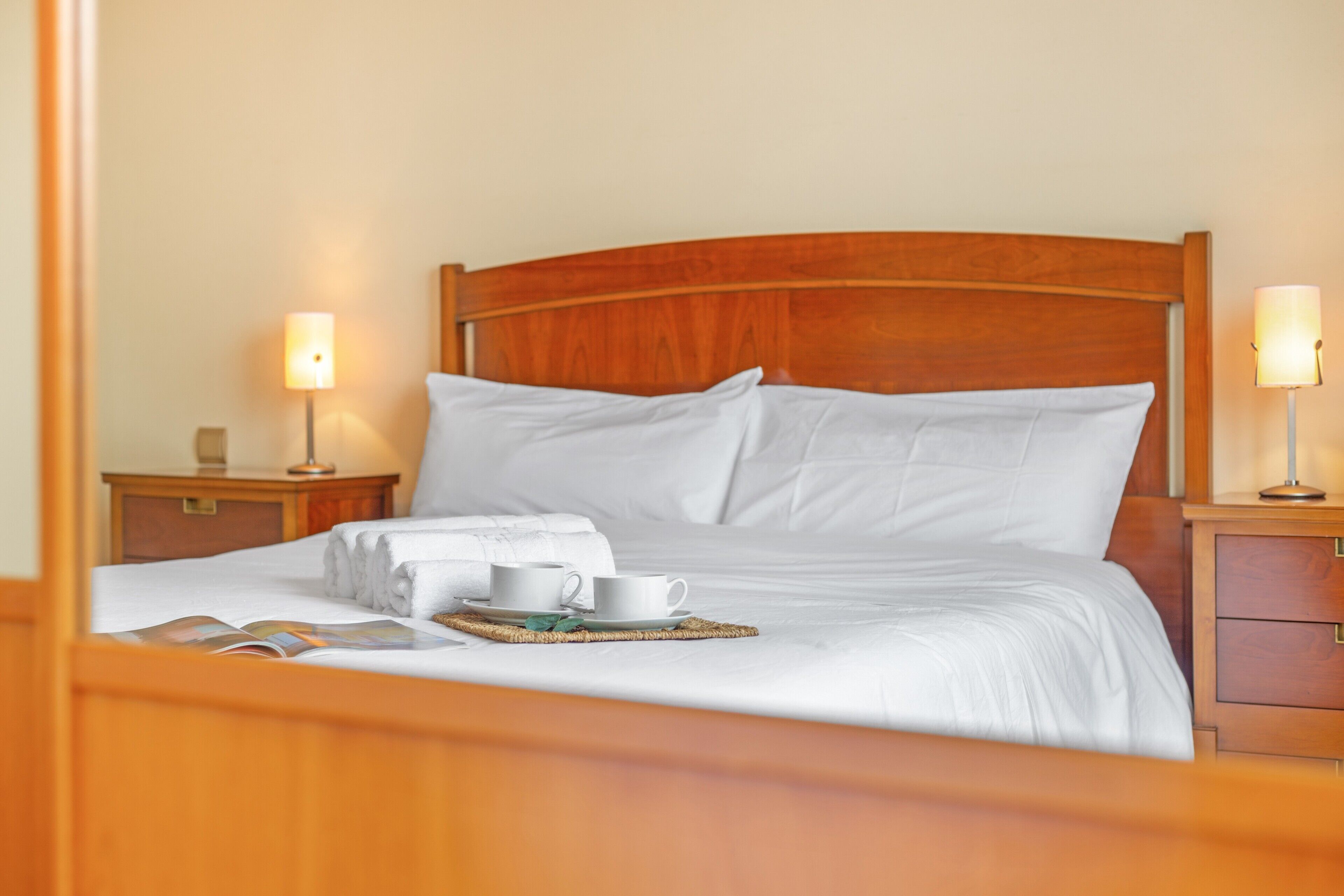 Una cama bien hecha con sábanas blancas, dos mesitas de noche con lámparas y una bandeja con un libro y dos tazas.