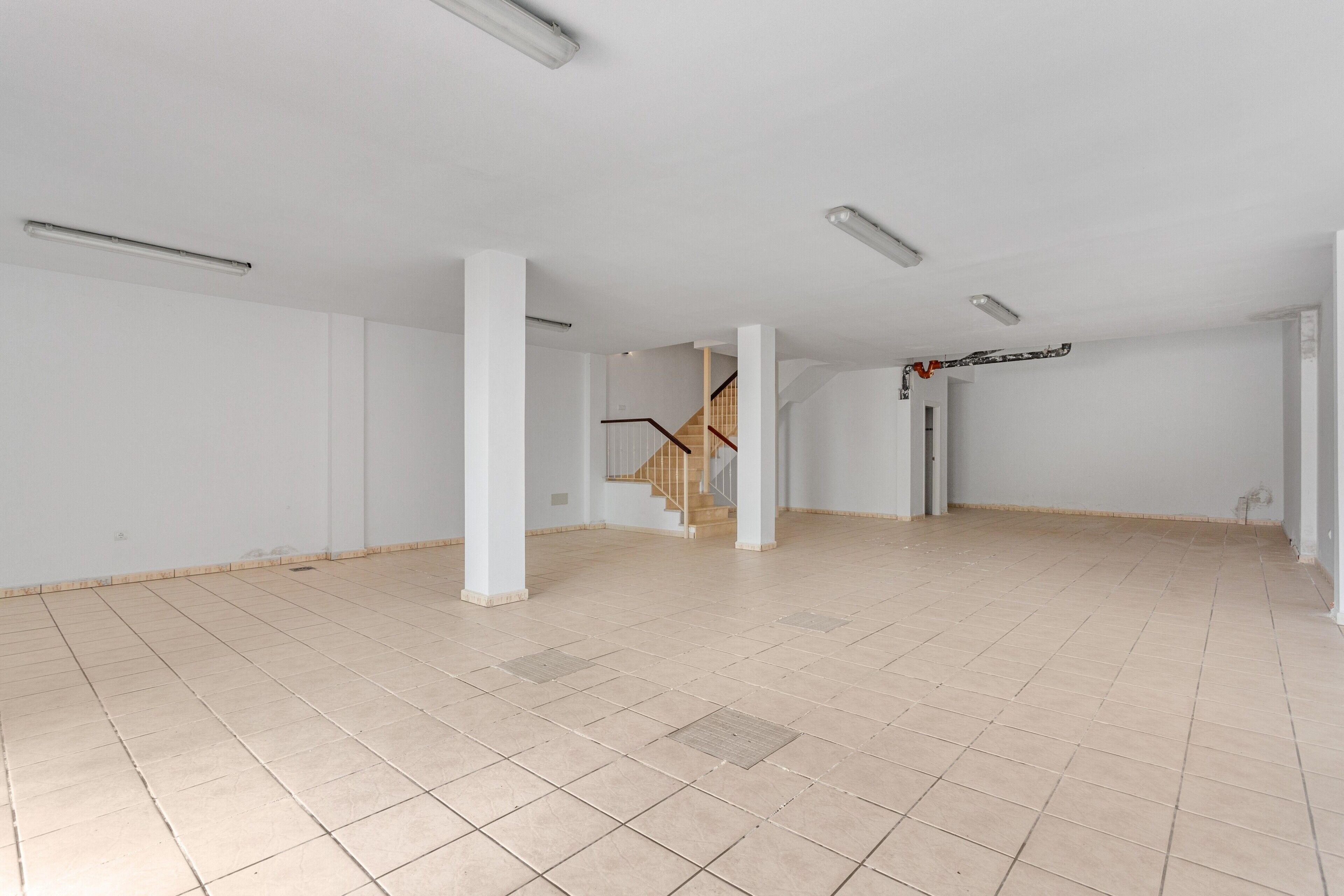 A spacious room with a tiled floor, white walls, and a wooden staircase.