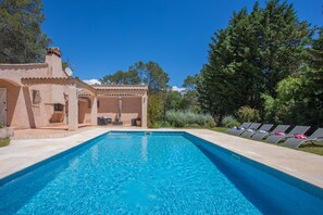 Ein klares, blaues Schwimmbecken mit Liegestühlen, einer überdachten Terrasse und einem rustikalen Gebäude mit Satellitenschüssel.
