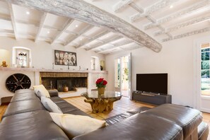 Een woonkamer met een grote leren bank, een ronde glazen salontafel, een open haard, een televisie en een rustieke plafond.